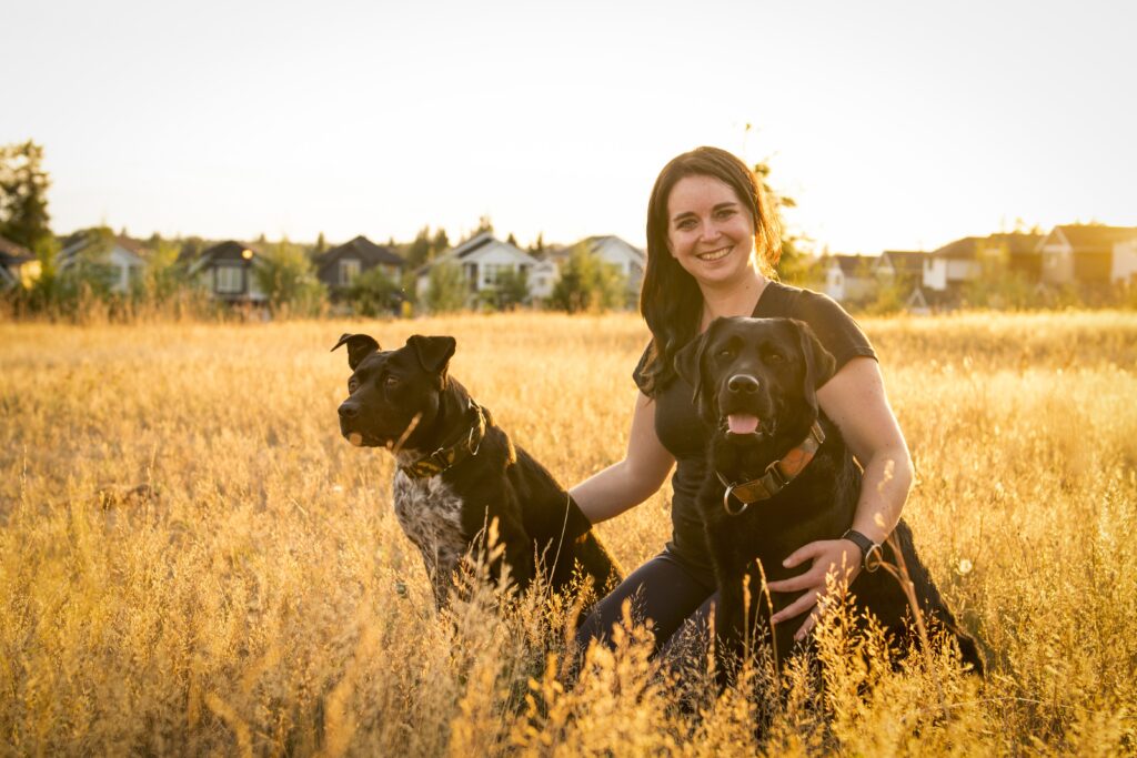 AnimalKind accredited trainer Amy Atkinson is kneeing in an open field looking at the camera and holding her dogs Staffy mix Midna and black Lab Archer 